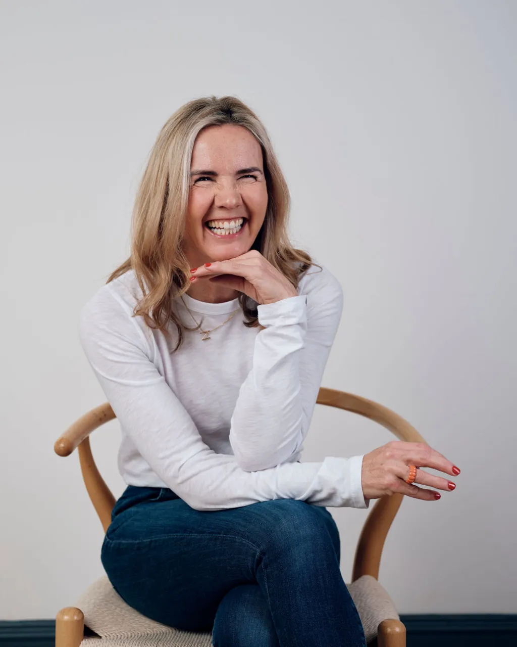 The coach laughs in a wooden chair, chin resting on her hand.